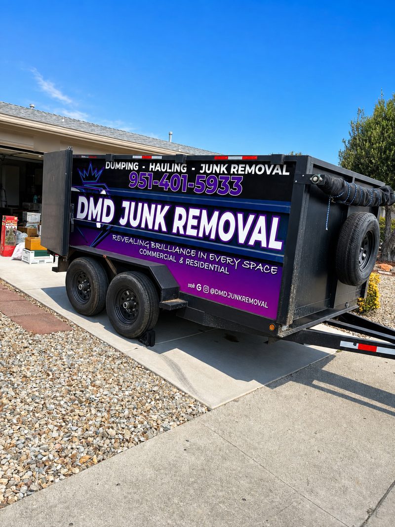 A DMD Junk Removal dump trailer parked on a residential driveway beside an open garage with boxes staged inside