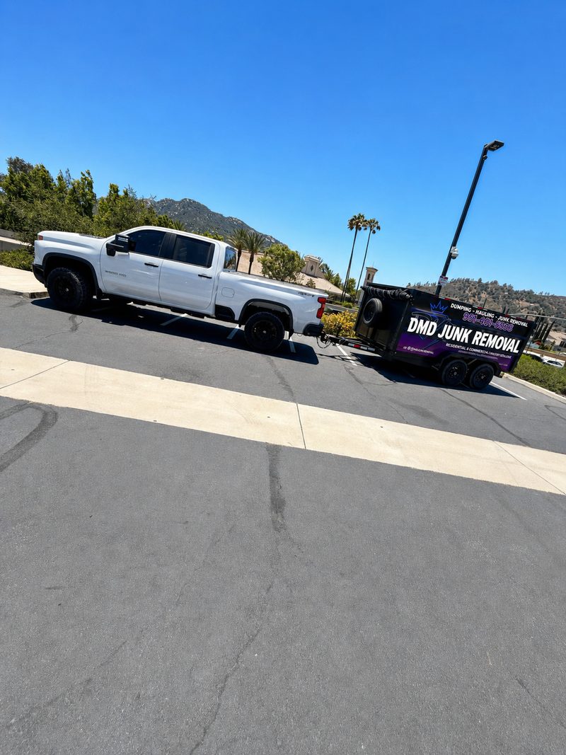 A white pickup towing a DMD Junk Removal trailer across a wide parking lot, hills and palm trees behind