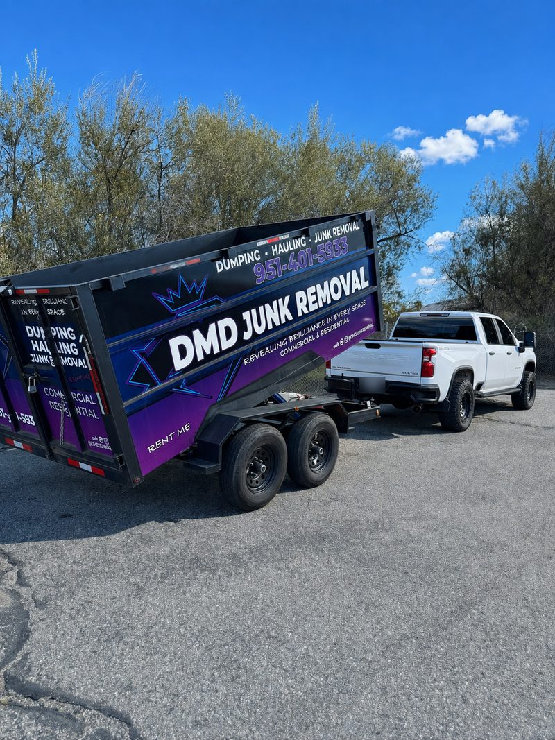A DMD Junk Removal dump trailer seen from the rear three-quarter, hitched to a white pickup under a clouded sky