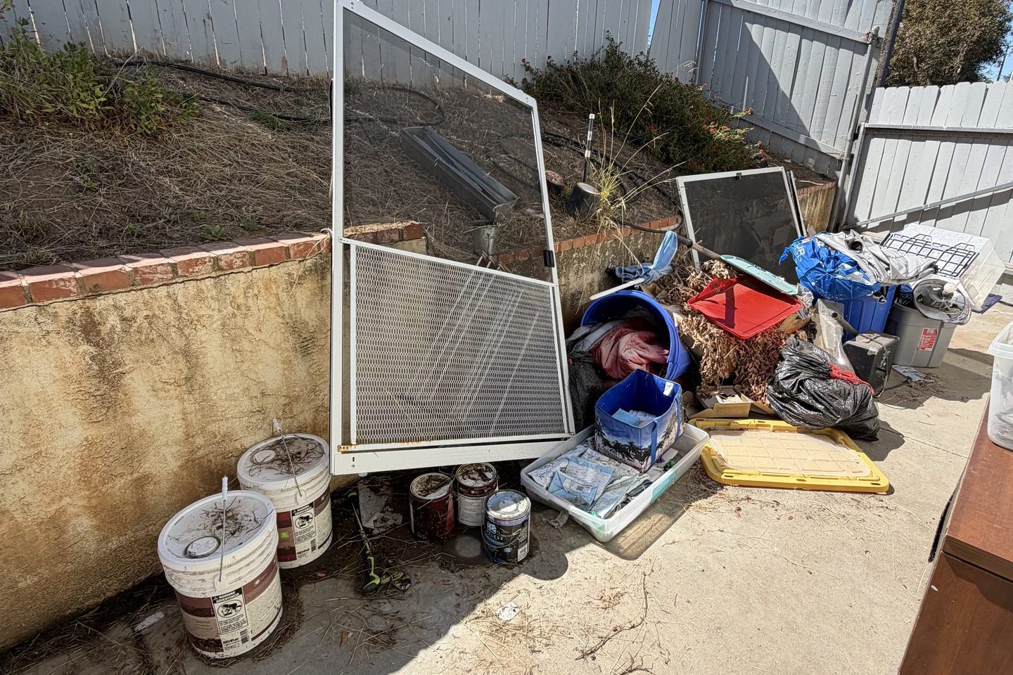 Household items cleared from a property and staged outside for haul-off — a screen door, paint buckets, bins and bagged debris