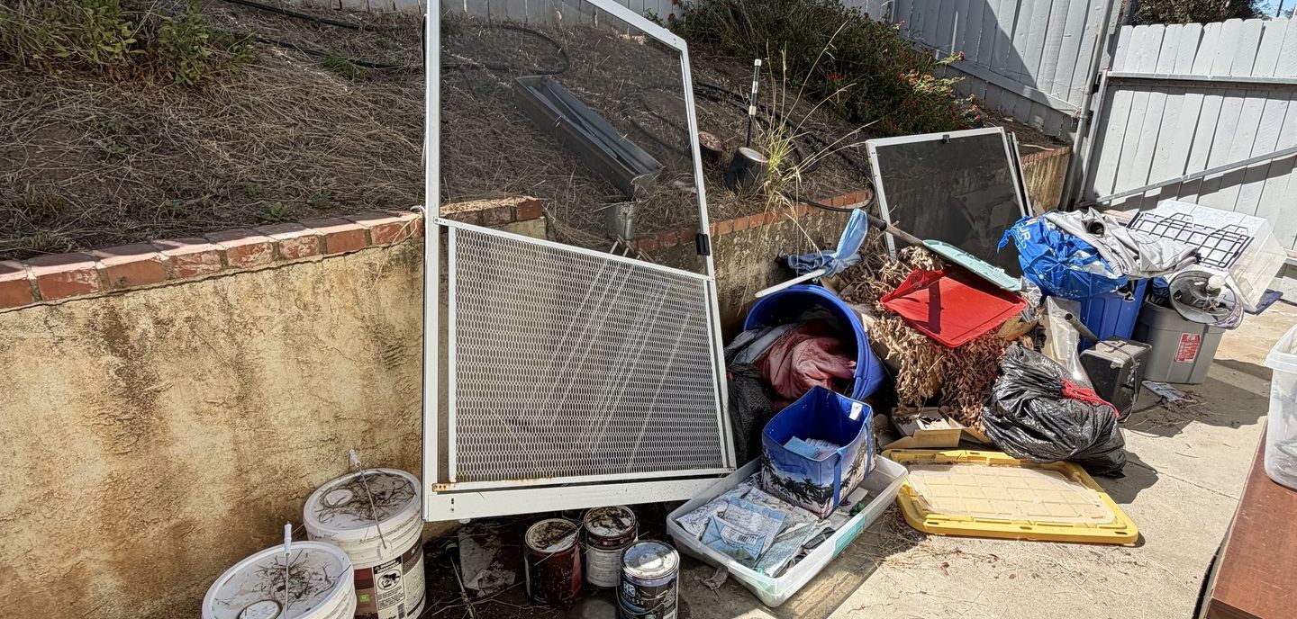 Household items cleared from a property and staged outside for haul-off — a screen door, paint buckets, bins and bagged debris