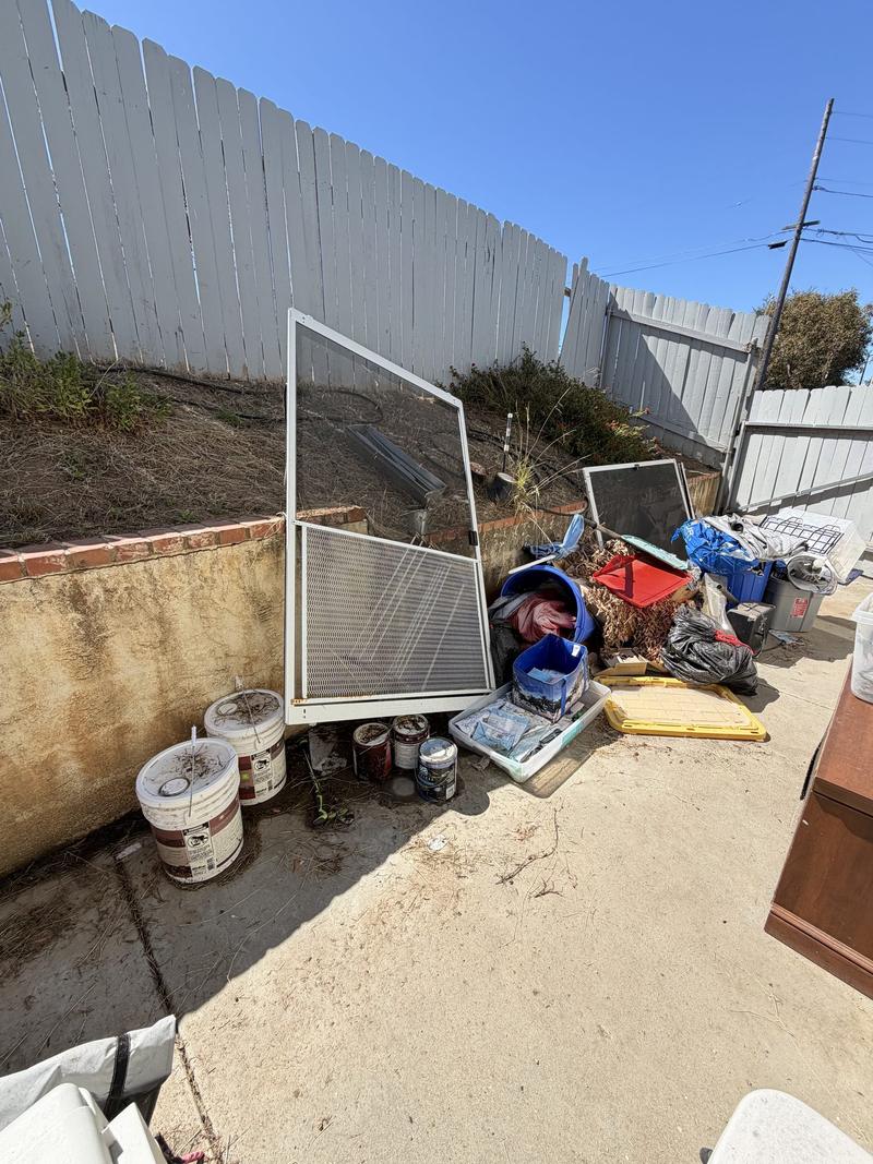 A side yard piled with an old screen door, paint buckets, bins and bagged debris before a DMD haul